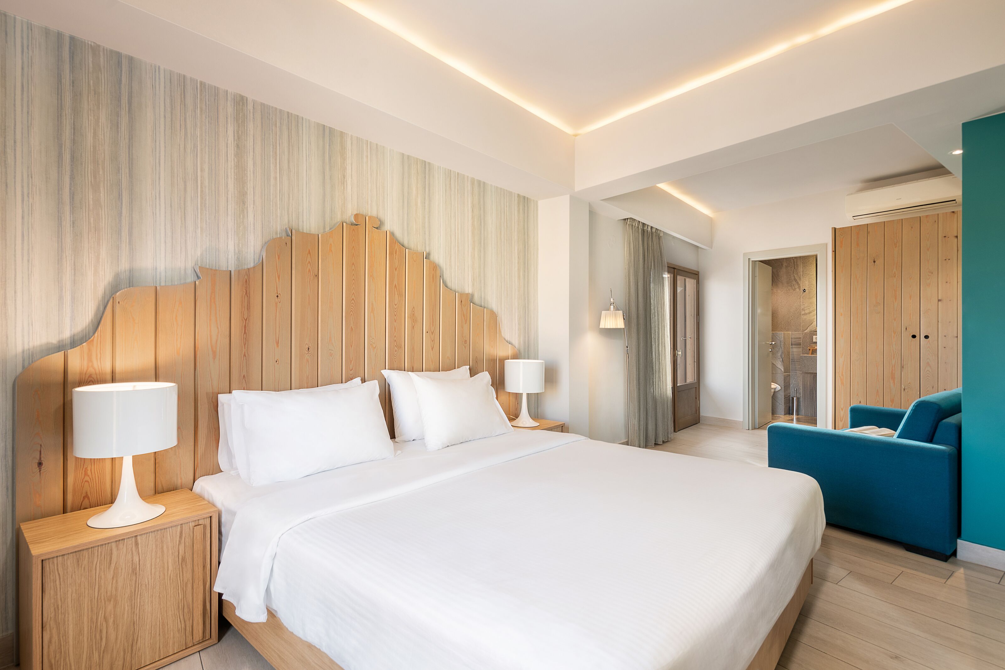 Een moderne hotelkamer met een groot bed, een houten hoofdbord, twee nachtkastjes met lampen, een blauwe fauteuil en uitzicht op de badkamer.
