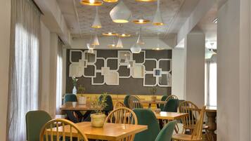 A restaurant interior with wooden tables and chairs, hanging pendant lights, and a geometric wall design.