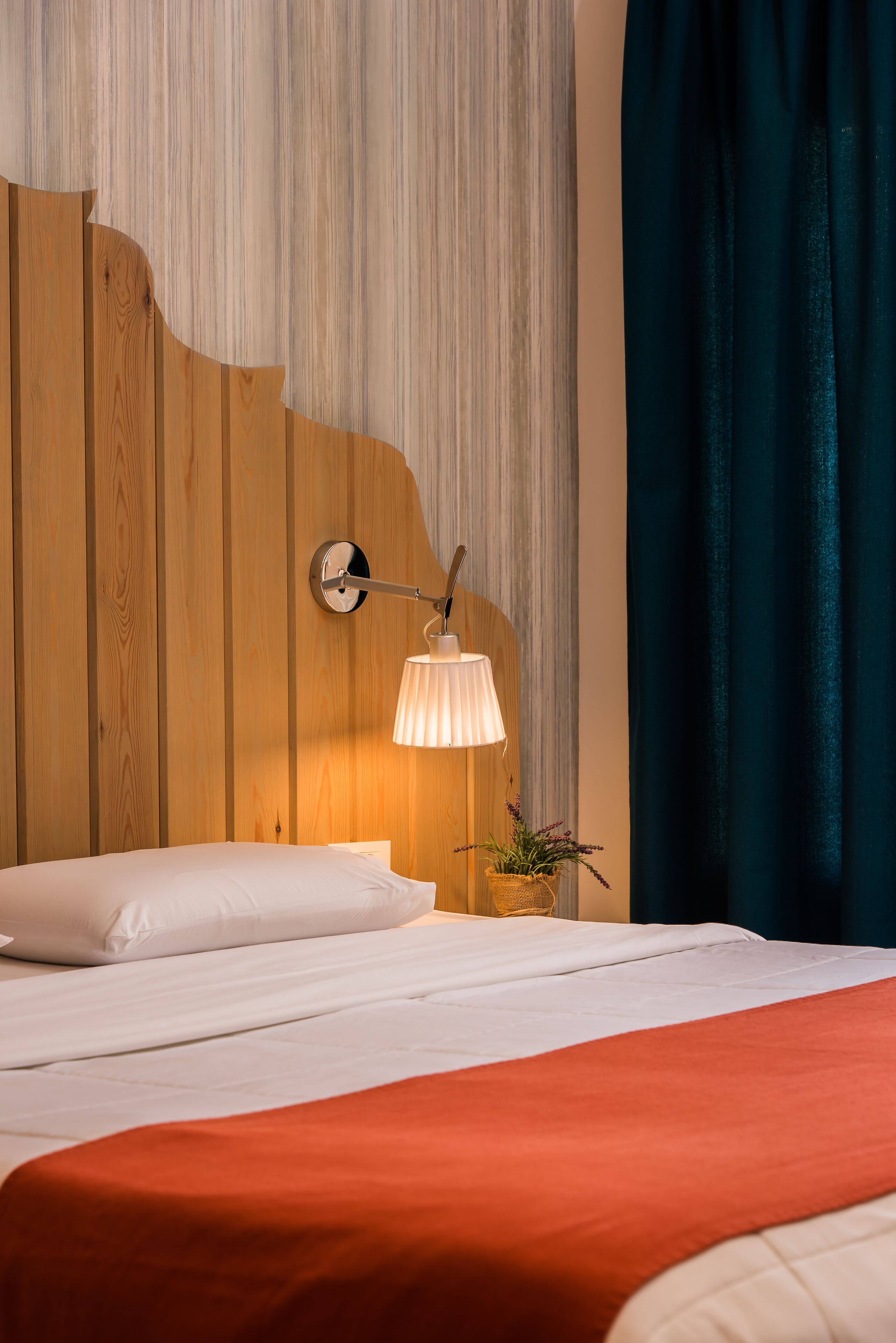 A neatly made bed with white linens and a red accent, a wall-mounted lamp, and a small potted plant on a wooden headboard.