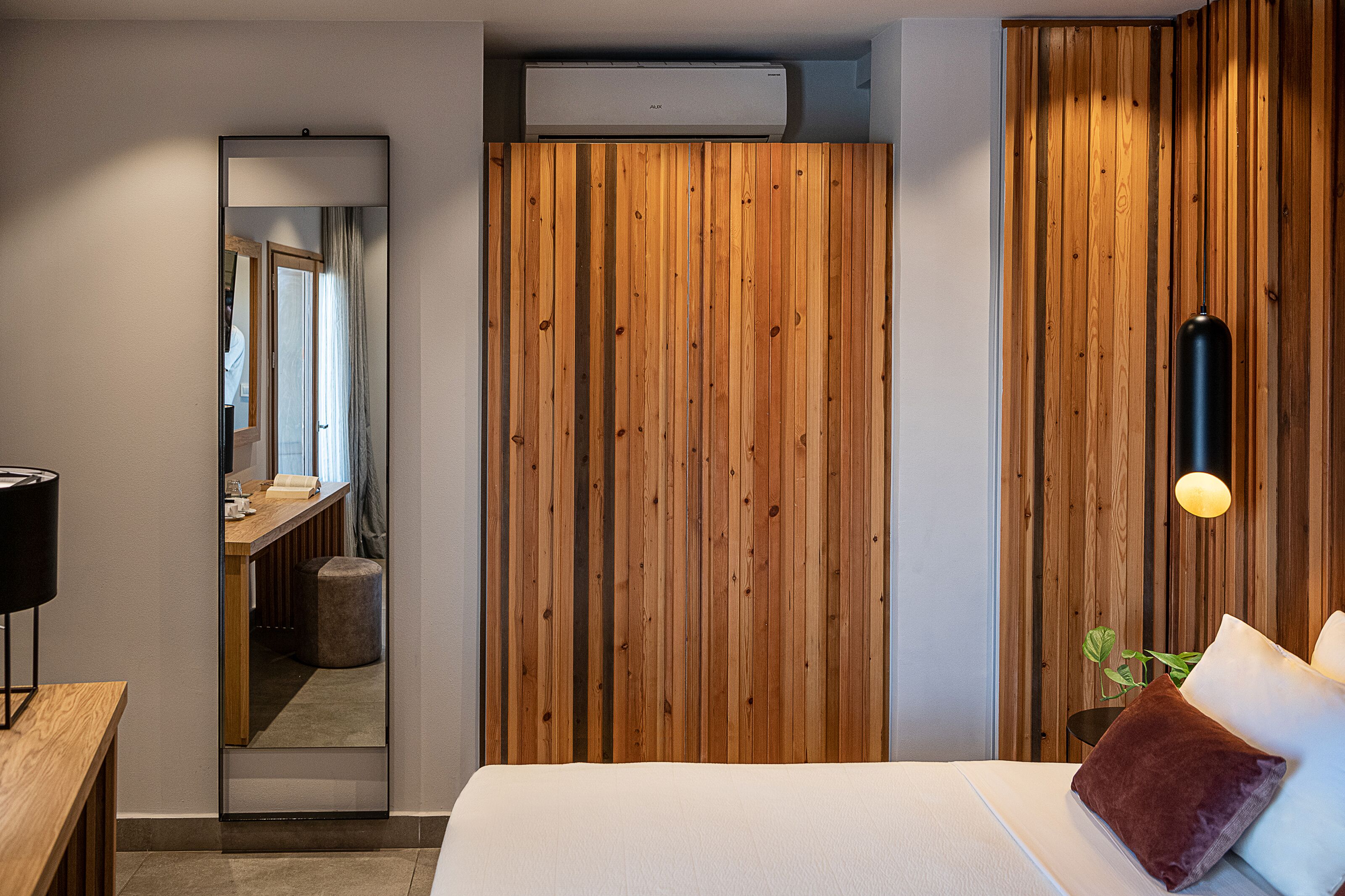 A modern hotel room with a wooden panel wall, a large mirror, a bed with pillows, a bedside table, and a hanging light fixture.