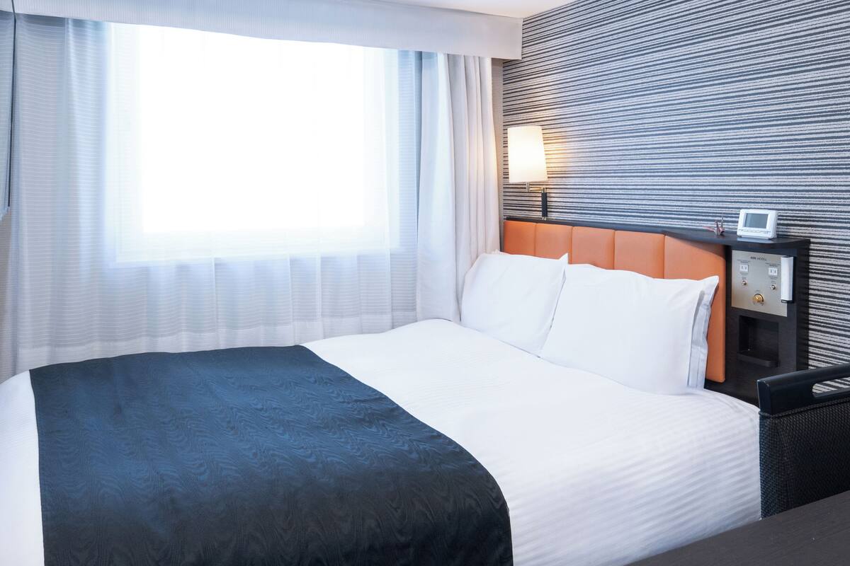 Quarto de hotel com uma cama grande, mesa de cabeceira com abajur e painel de controle fixado na parede.