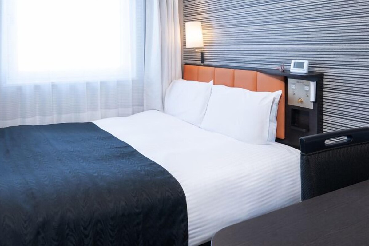 A hotel room with a large bed, a nightstand with a lamp, and a view of the outside through sheer curtains.