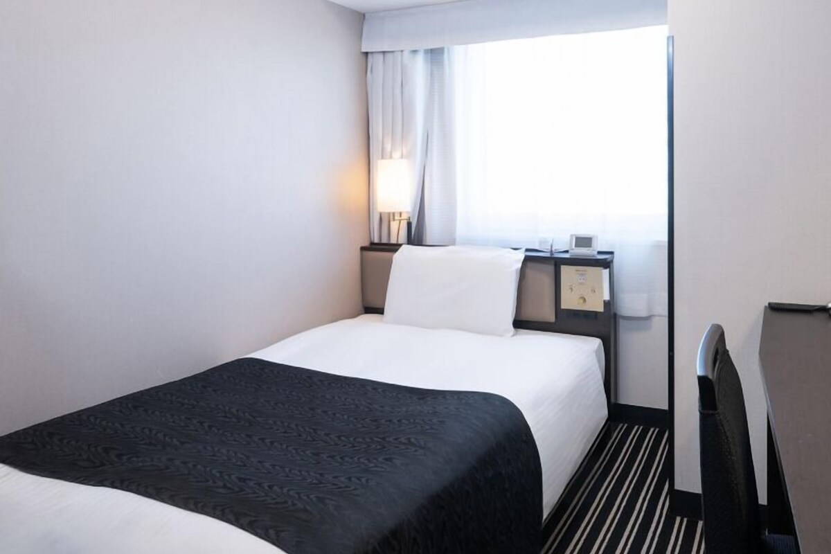 A hotel room with a bed, a bedside table with a lamp, a nightstand with a clock, and a window with curtains.