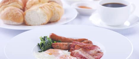 A breakfast plate with eggs, bacon, sausages, and greens, accompanied by croissants and a cup of coffee on a white table.