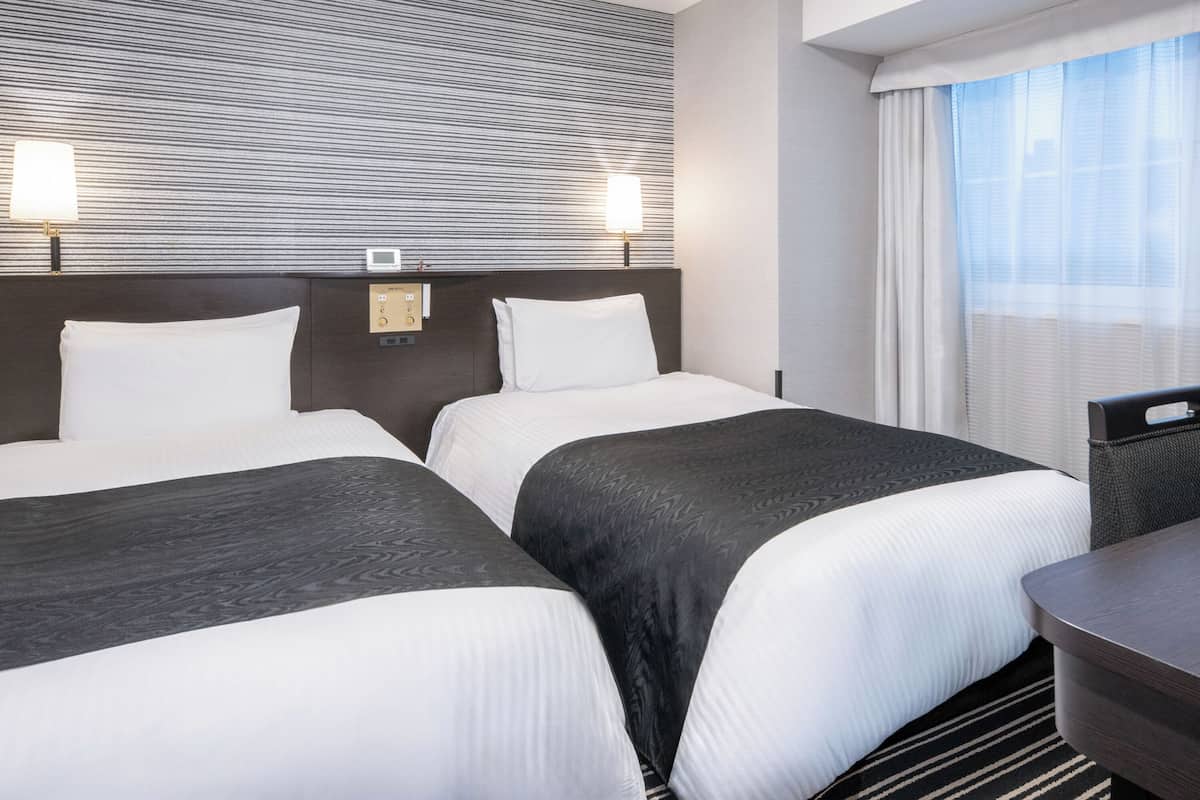 Quarto de hotel com duas camas, cabeceira com painel de controle, abajures, uma escrivaninha e uma janela com cortinas translúcidas.