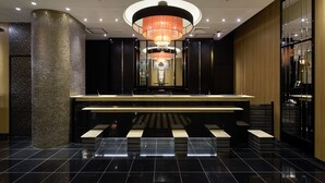 Um lobby moderno de hotel, com um balcão de recepção central, um grande candelabro e um chão de azulejos preto e branco polido.