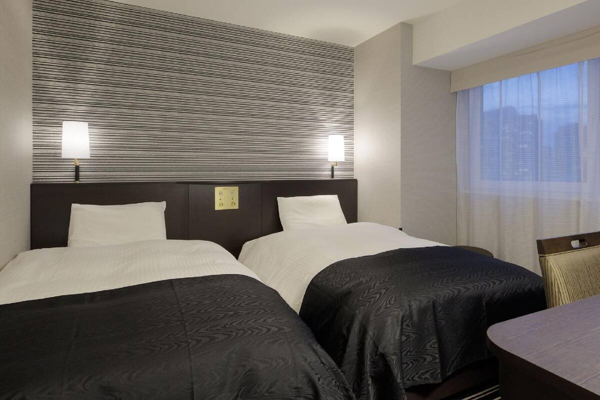 Une chambre d’hôtel avec deux lits, un bureau et une fenêtre avec des stores.