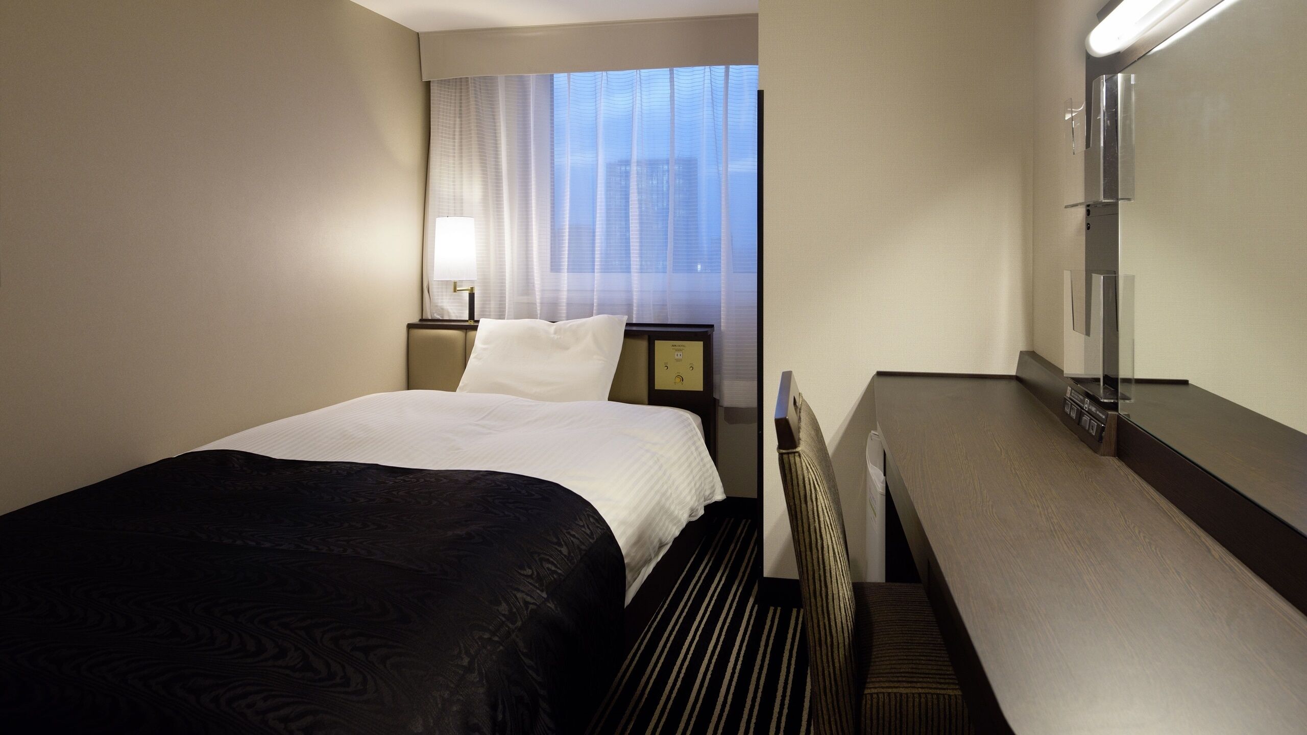 Een hotelkamer met een bed, een bureau, een lamp, een nachtkastje en een raam met gordijnen.