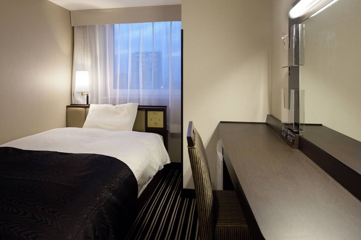 Une chambre d’hôtel avec un lit, un bureau, une lampe, une table de chevet et une fenêtre avec des rideaux.