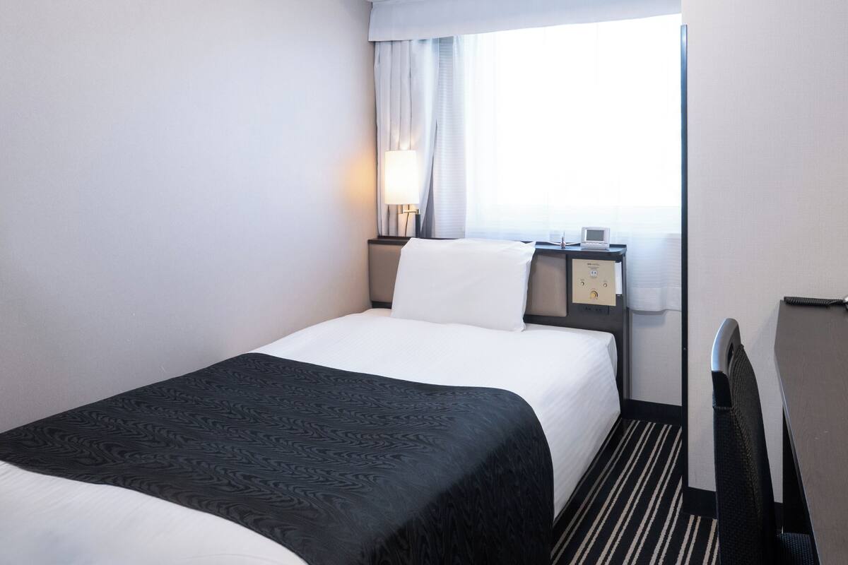 Quarto de hotel com cama, mesa de cabeceira, abajur, painel de controle e janela com cortinas.