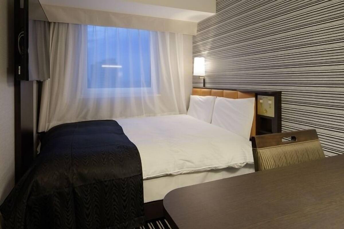 Une chambre d’hôtel avec un lit, une table de chevet, une lampe et une fenêtre avec des rideaux.