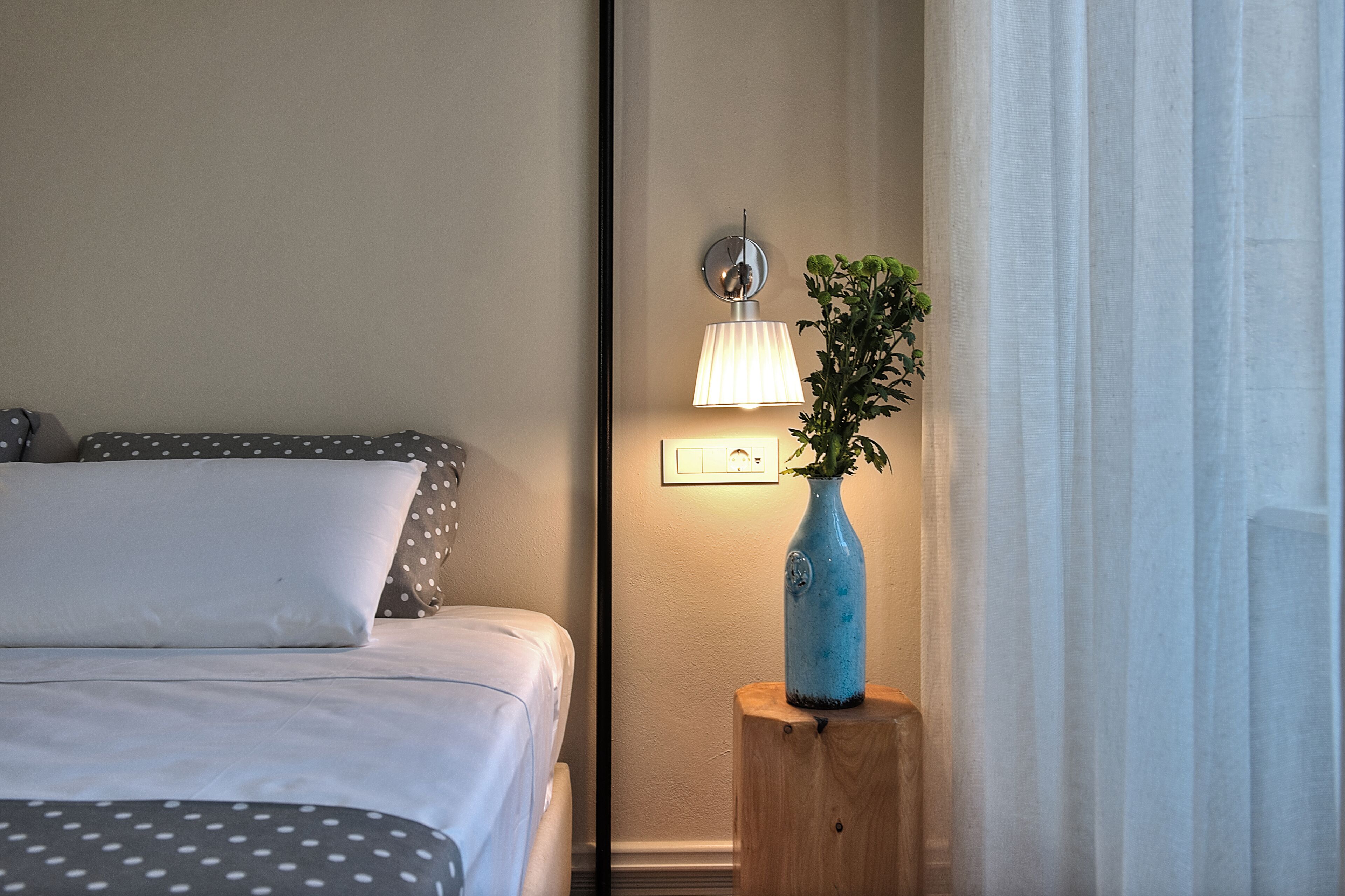 Una cama bien hecha con ropa de cama gris y blanca con lunares, un jarrón azul con una planta en una mesita de noche de madera, y una lámpara fijada a la pared.