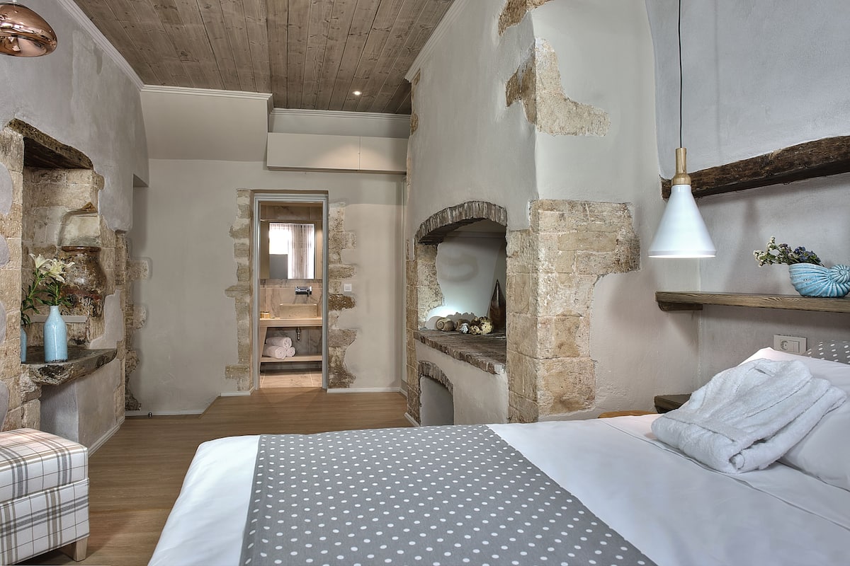 Une chambre avec des murs en briques apparentes, une cheminée en pierre, un lit avec une literie blanche et un luminaire suspendu.