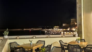 Un área de comedor al aire libre con mesas y sillas, con vista al frente marítimo, edificios y un puerto.