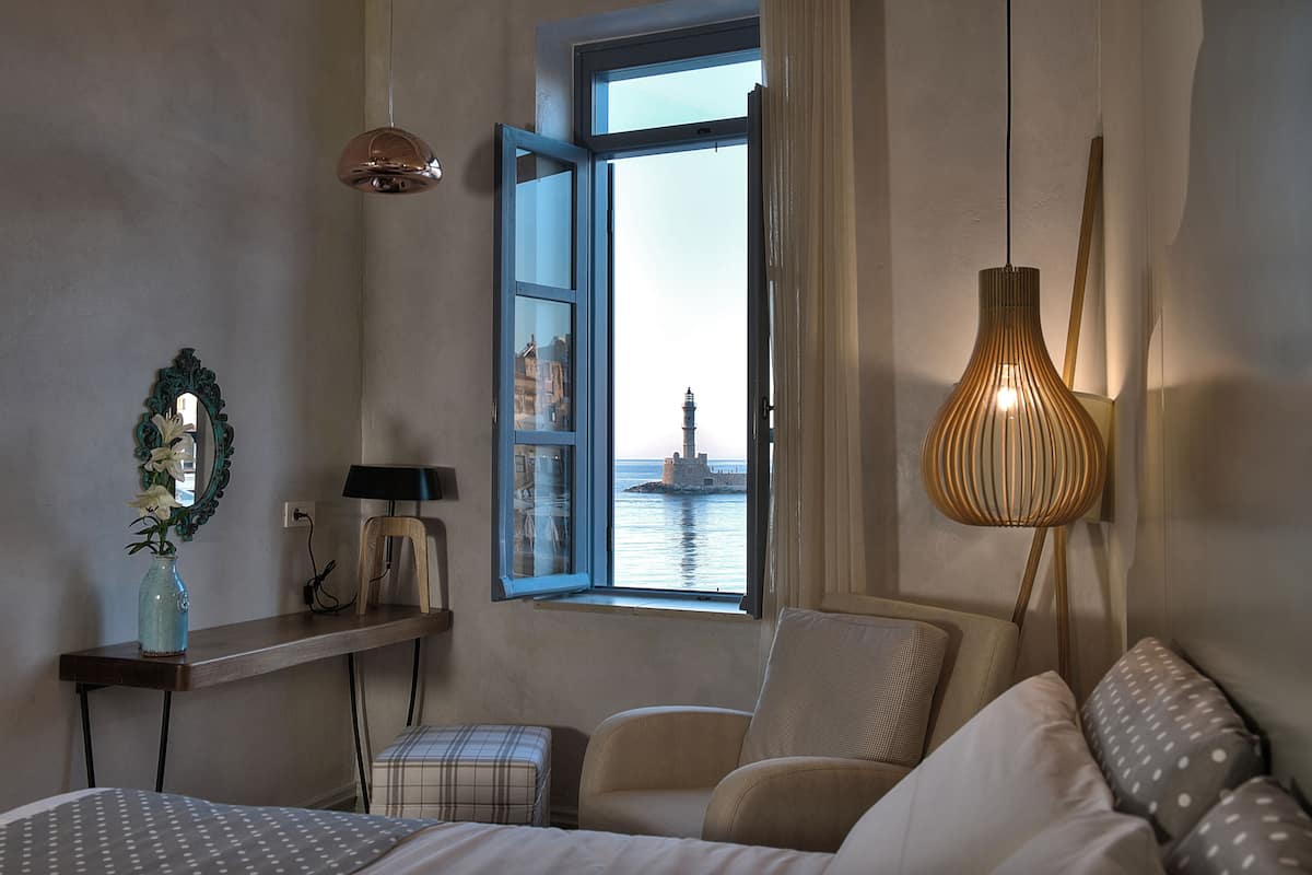 Une chambre d’hôtel avec un lit, une chaise, une petite table sur laquelle se trouve un vase, une lampe et une fenêtre donnant sur un phare.