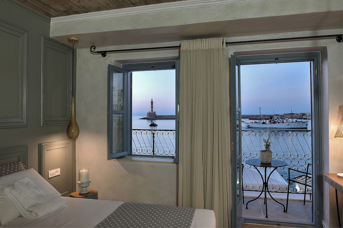 Une chambre d’hôtel avec un lit, une table de chevet avec une bougie, une fenêtre donnant sur un plan d’eau et une tour lointaine, et un balcon avec une table et une chaise.