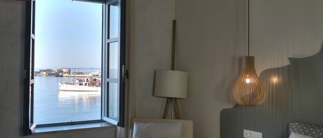 A hotel room with a large window offering a view of a harbor, a chair, a bedside table, and a lamp.