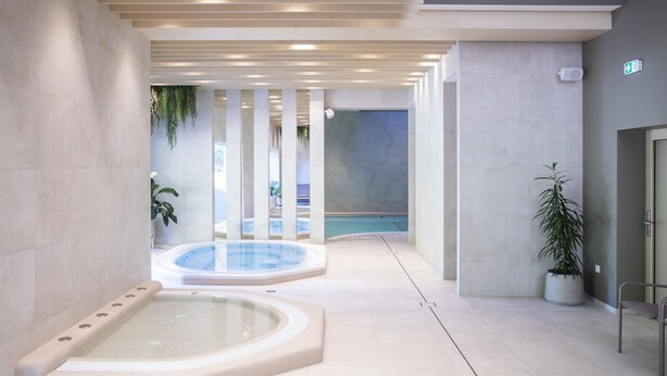 Un área interior de piscina moderna con dos jacuzzis, un espacio de descanso y vistas a una piscina exterior.