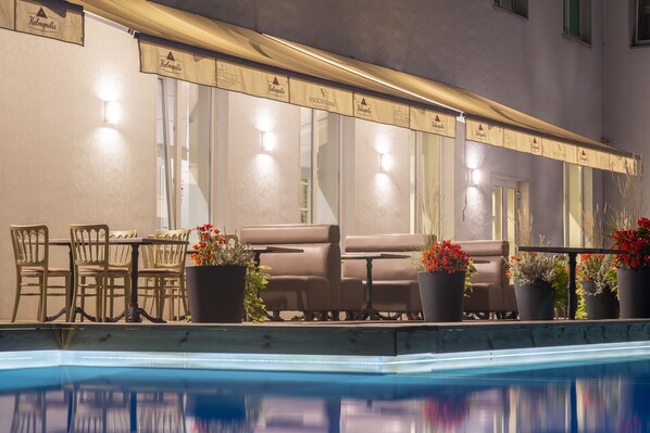 Un área de descanso junto a la piscina con mesa y sillas, plantas en macetas y un reflejo en el agua.