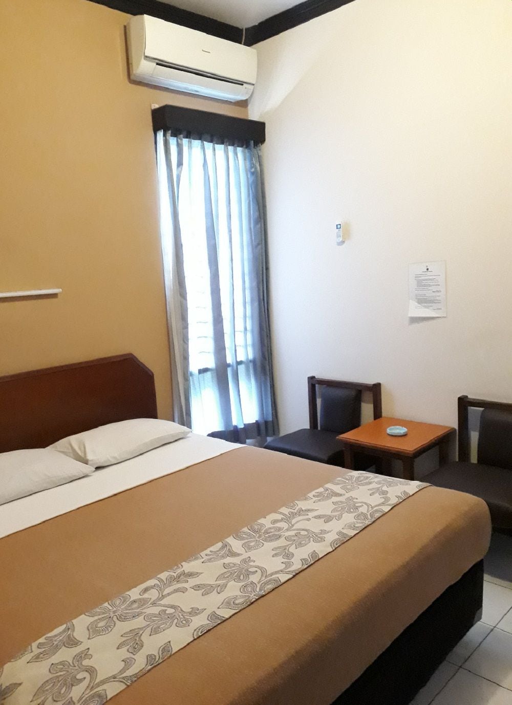 A hotel room with a bed, a small table, a chair, an air conditioning unit, and a window with curtains.