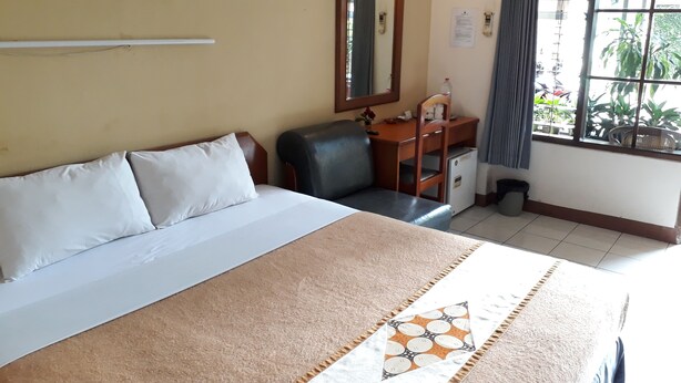 A hotel room with a bed, a desk, a chair, a small refrigerator, a window with curtains, and a mirror on the wall.