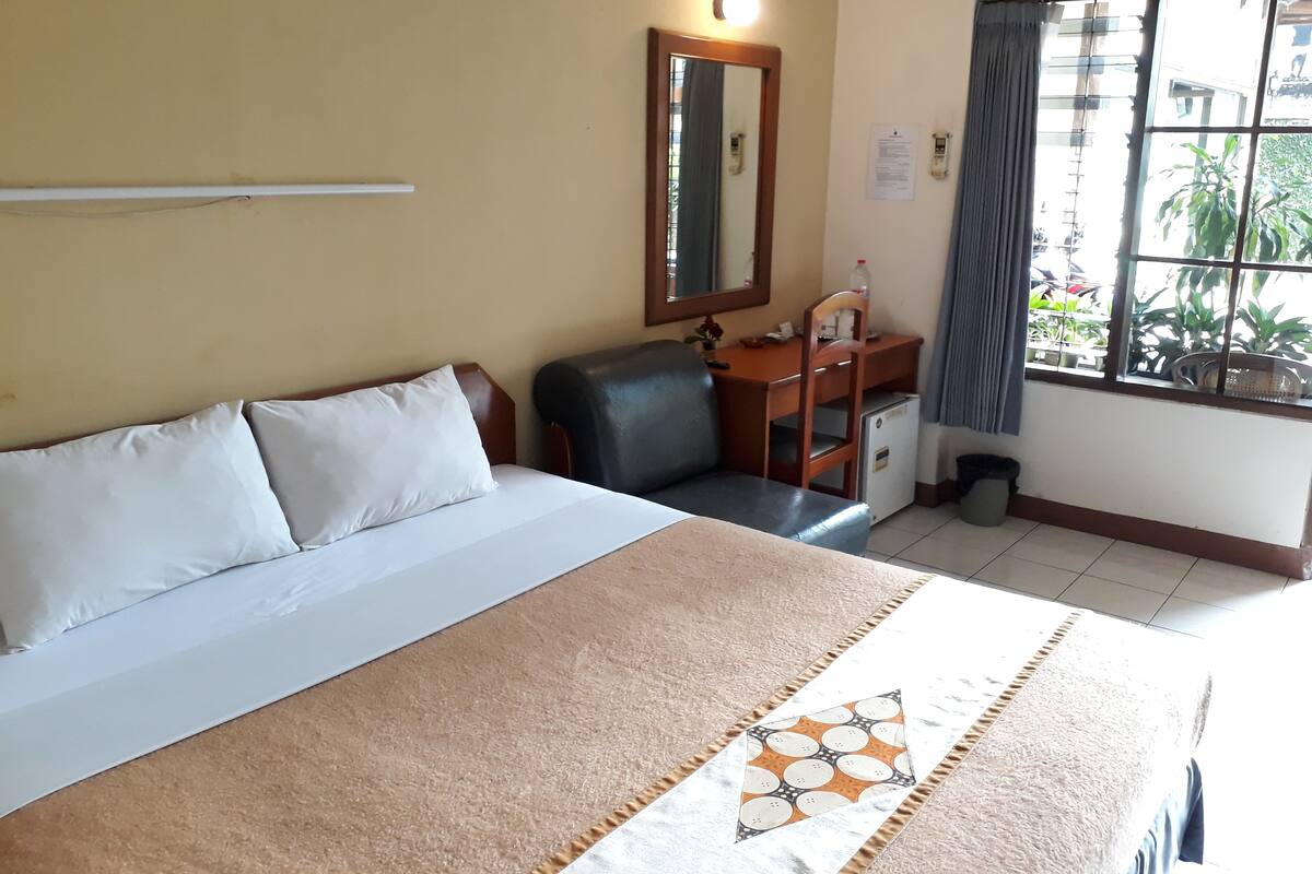 A hotel room with a bed, a desk, a chair, a small refrigerator, a window with curtains, and a mirror on the wall.
