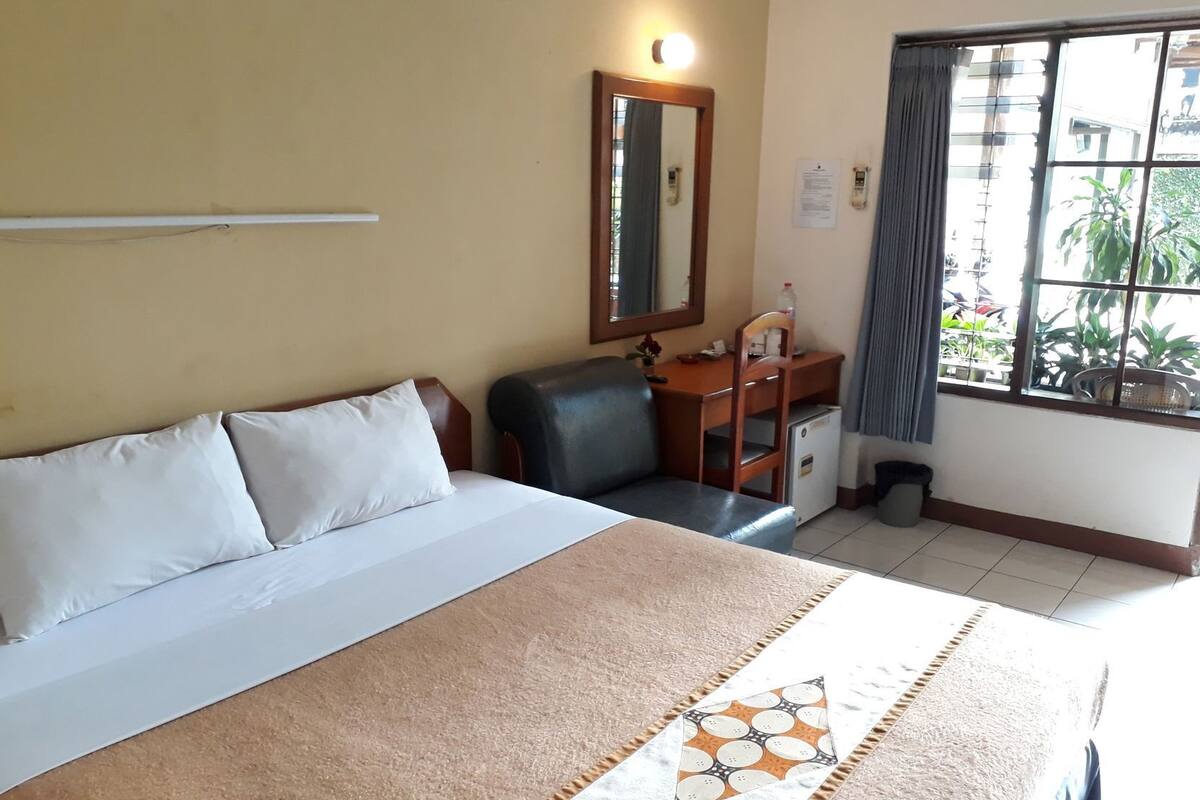 A hotel room with a bed, a desk, a chair, a small refrigerator, a window with curtains, and a mirror on the wall.