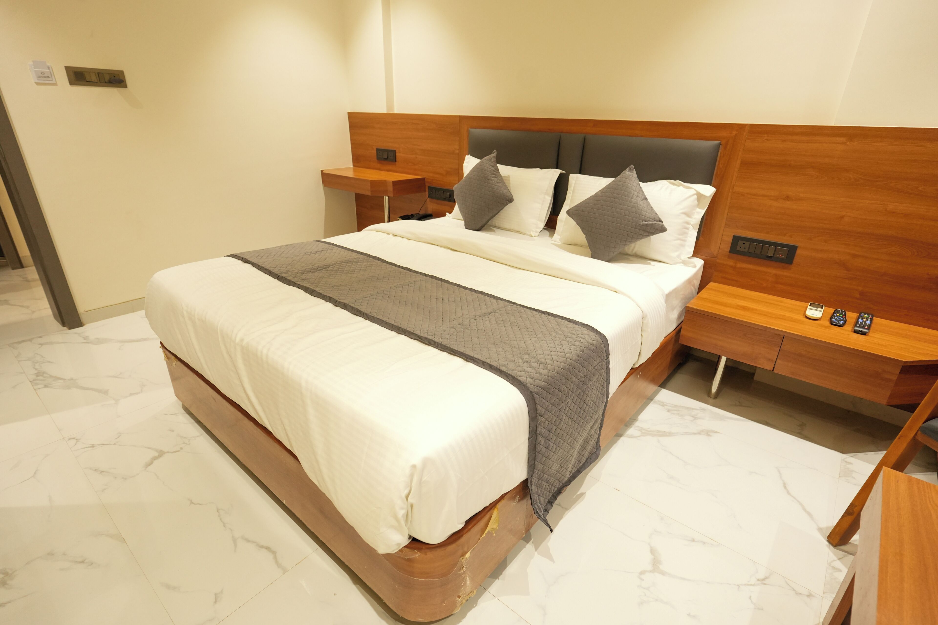 Una cama bien tendida con cabecera de madera, una mesita de noche con teléfono y control remoto, y un suelo con diseño de mármol.
