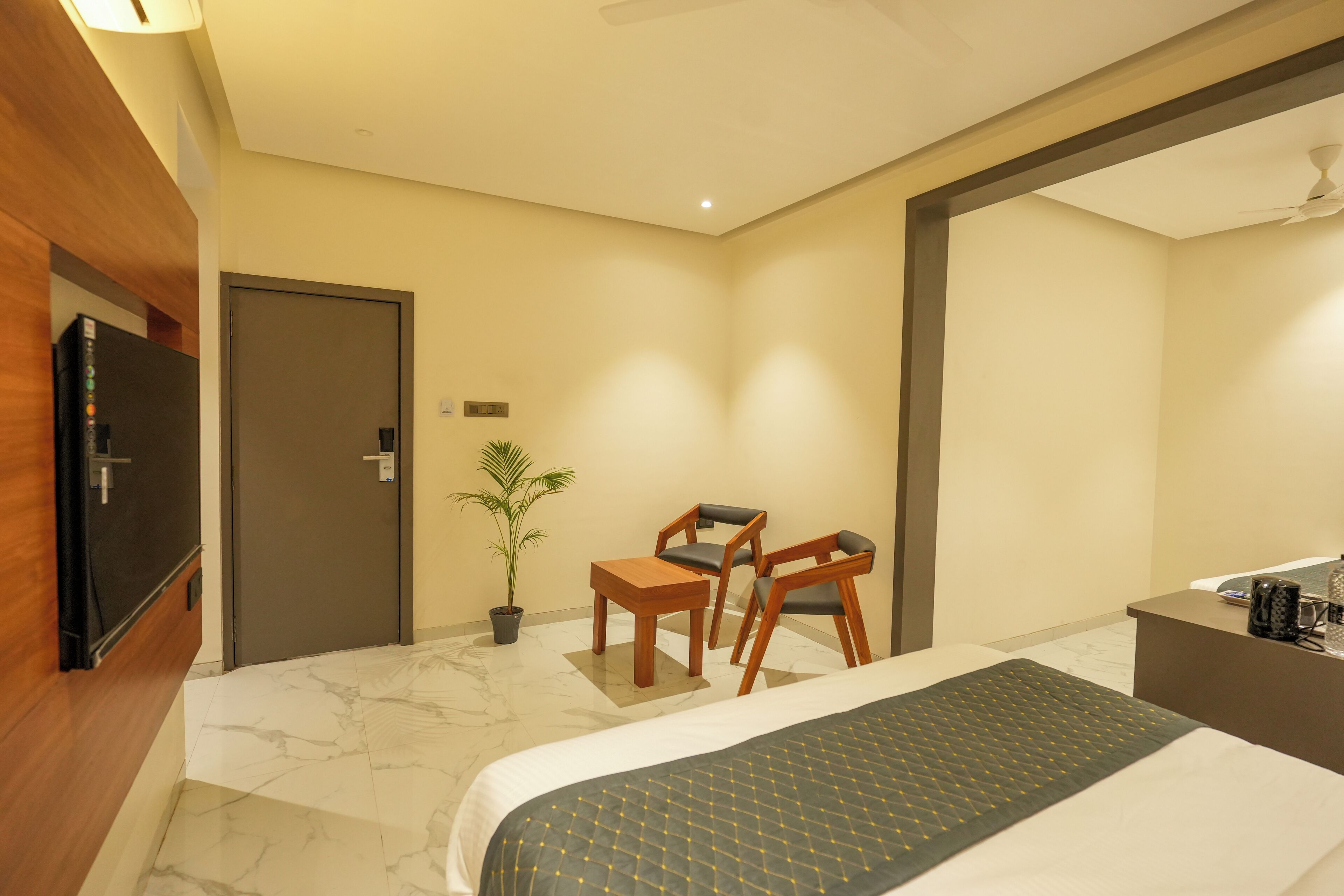 Habitación de hotel con cama, escritorio, dos sillas, televisor, planta en maceta y mostrador de recepción.