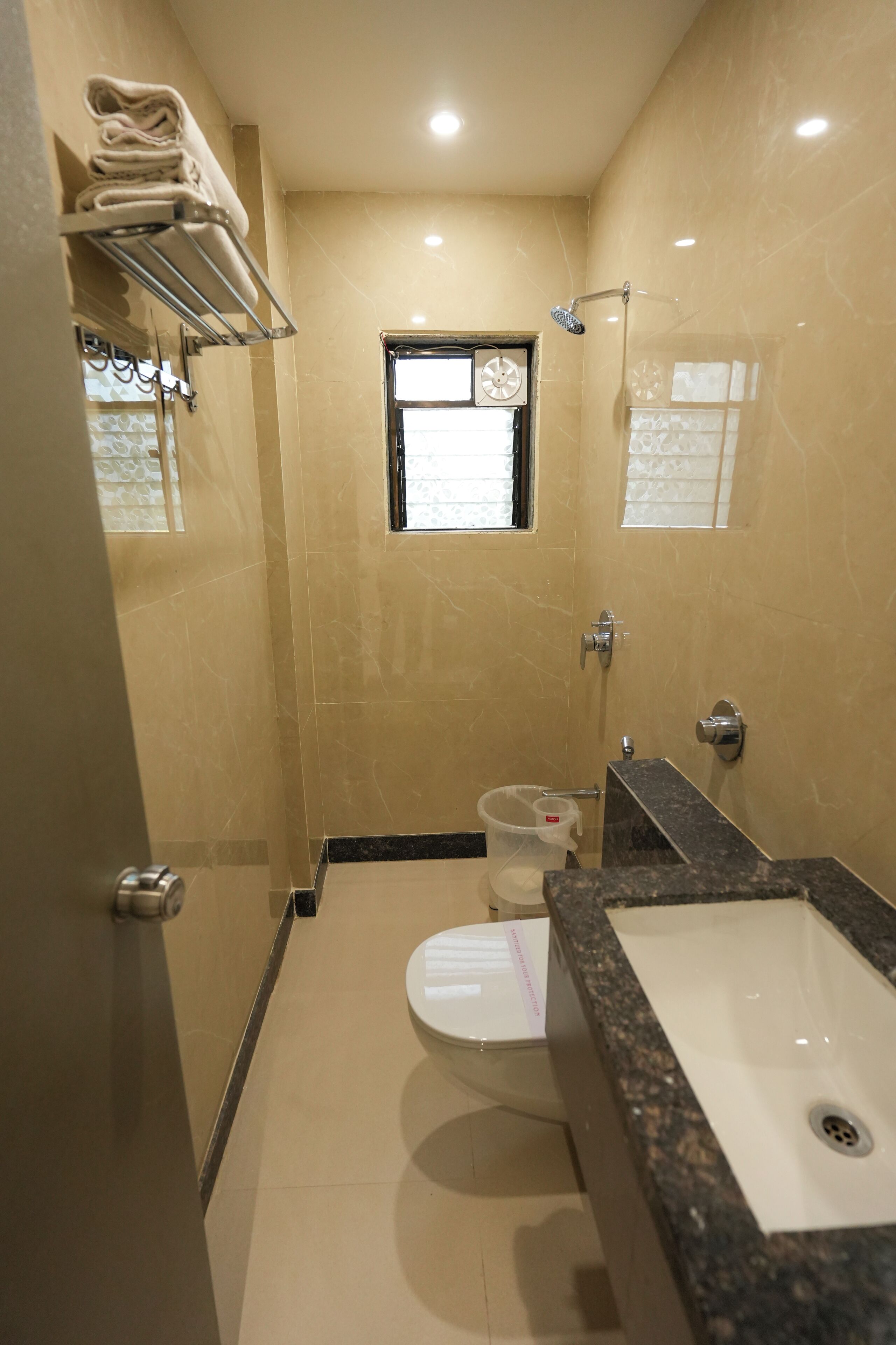 A bathroom with a toilet, sink, and shower. There is a window above the toilet and a shelf with towels.