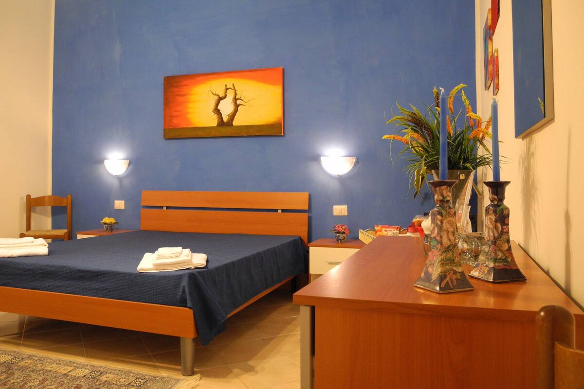 A hotel room with a bed, a desk, a chair, a nightstand, a wall-mounted lamp, a painting, and a vase with flowers.