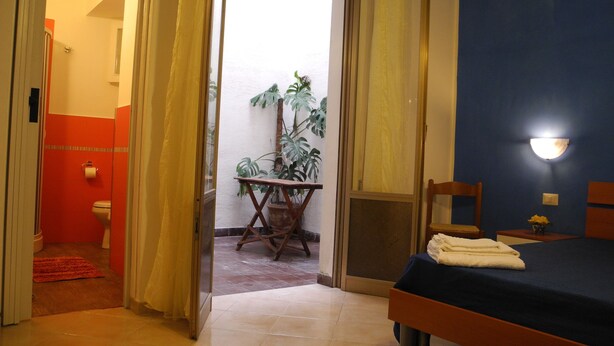 A bedroom with a bed, a bedside table, a nightstand, a potted plant, and a bathroom visible through an open door. - Melone Giallo (Paceco)
