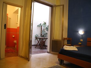 Quarto com cama, mesa de cabeceira, planta em vaso e banheiro visível através de uma porta aberta.