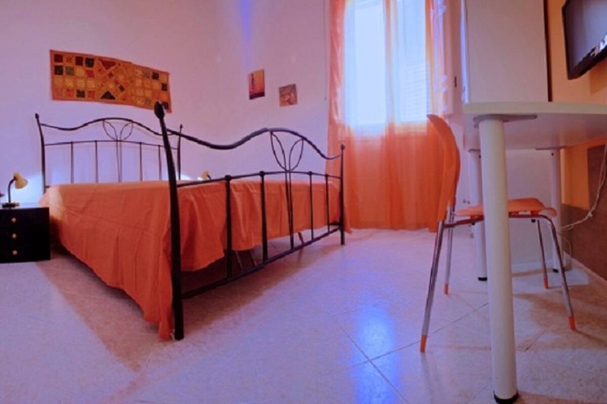 A bedroom with a bed, a nightstand, a chair, a television, and a window with curtains.