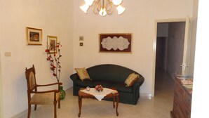 A living room with a dark green sofa, a wooden chair, a small table with a vase, and a chandelier. - Melone Giallo (Paceco)