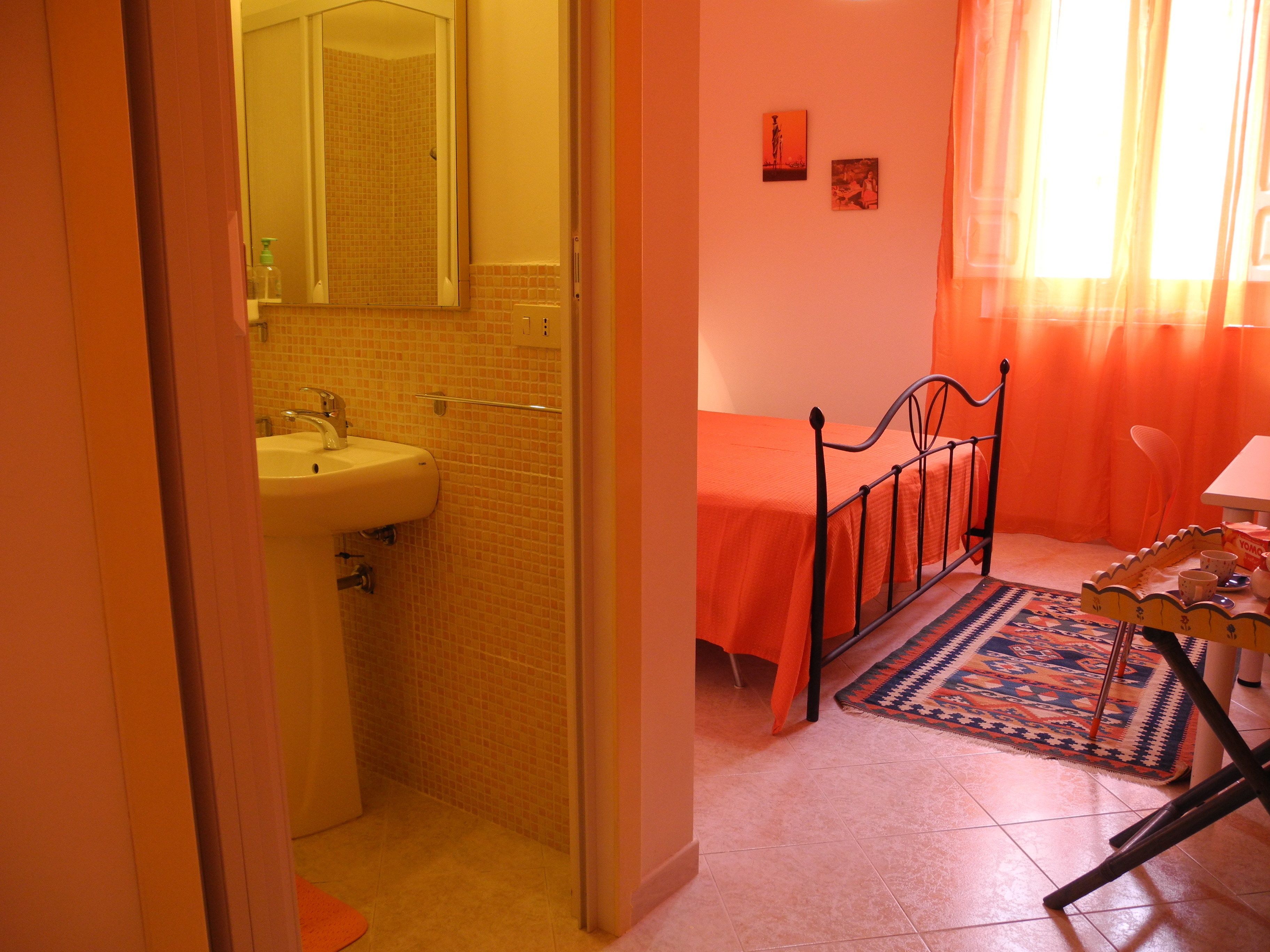 Un bagno con lavandino e specchio, un letto con copripiumino arancione e un tavolino con tazza e piattino.