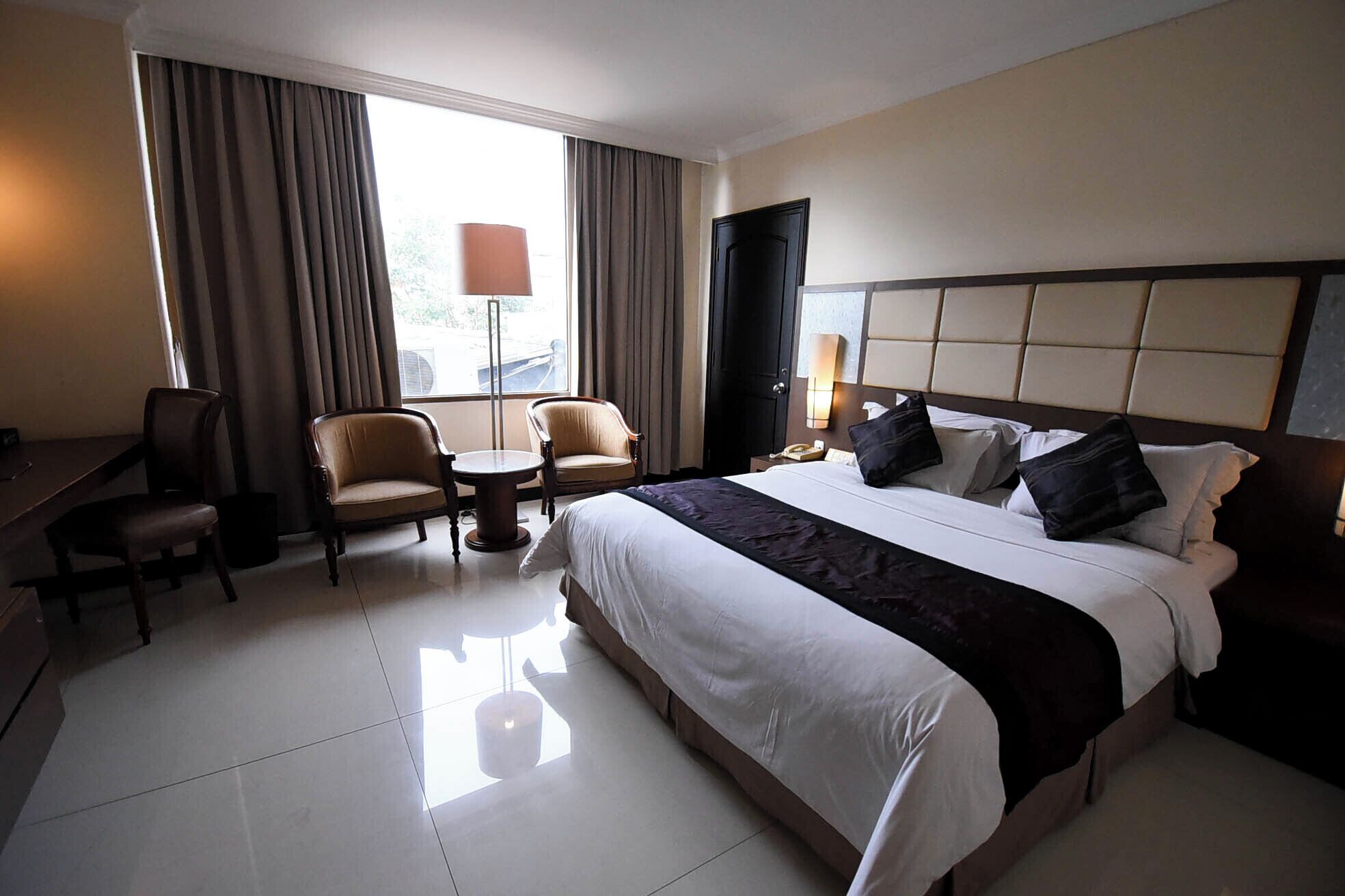 Ein Hotelzimmer mit einem großen Bett, zwei Sesseln, einem kleinen Tisch, einem Schreibtisch und einem Fenster mit Vorhängen.
