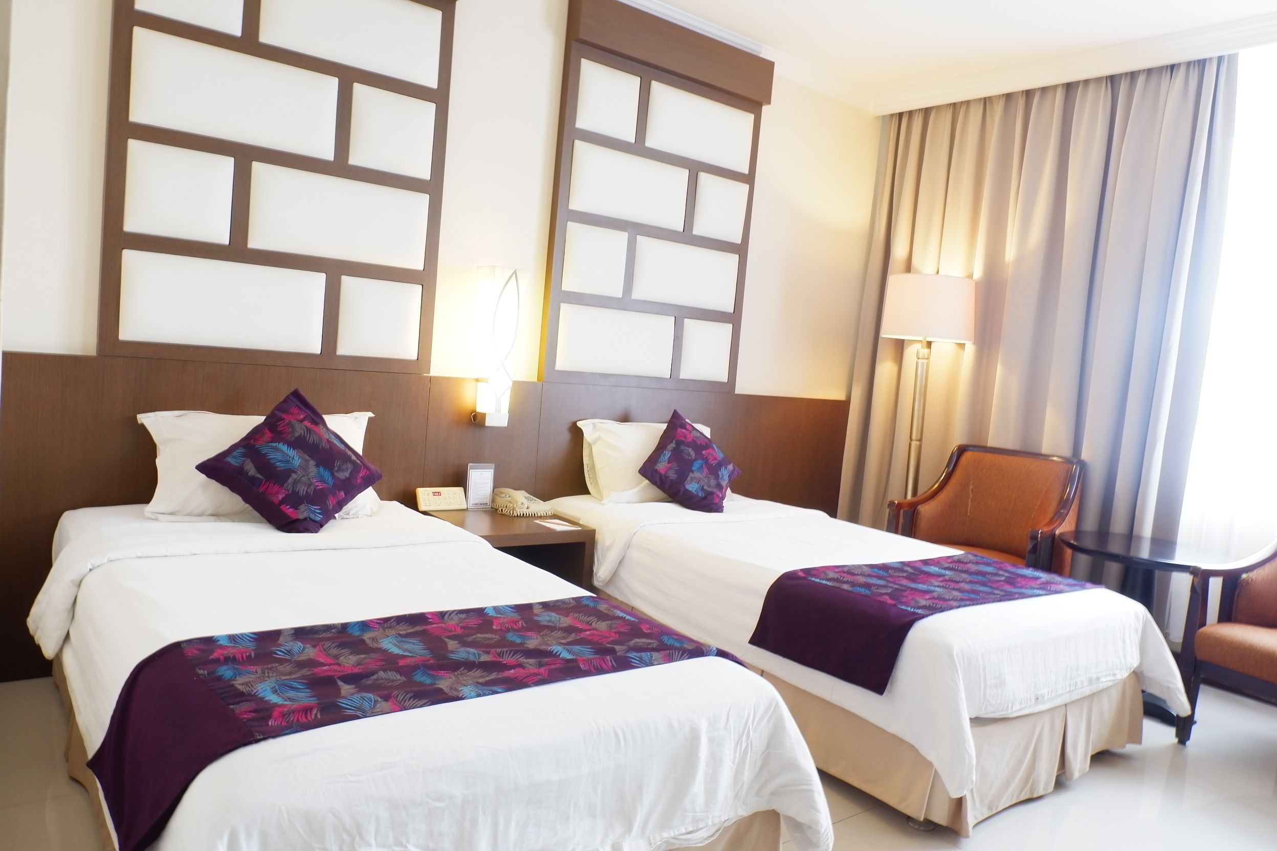 Ein Hotelzimmer mit zwei Betten, einem Schreibtisch, einem Stuhl und einem großen Fenster mit Vorhängen.