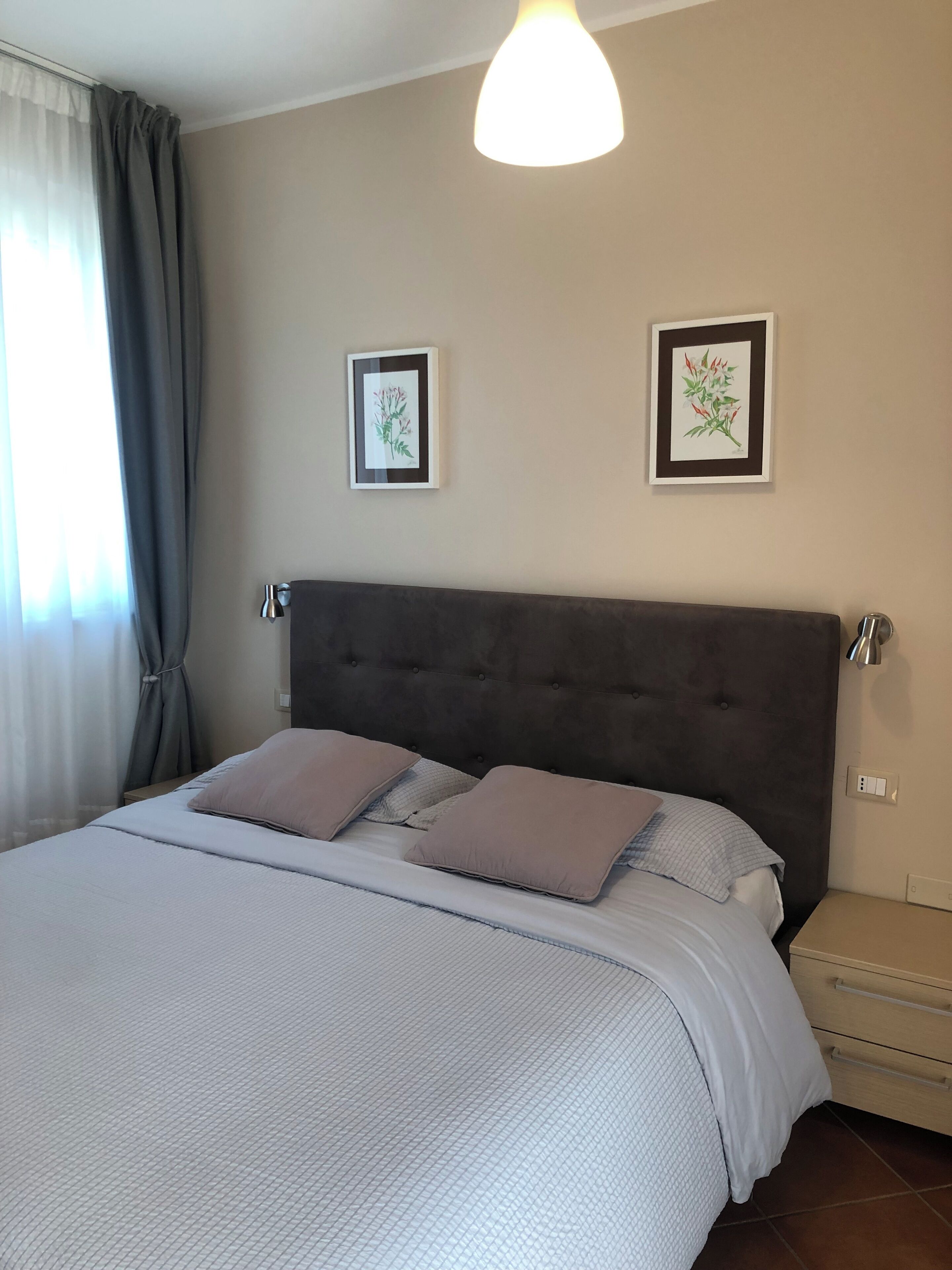 Una cama bien hecha con cabecera gris, dos mesitas de noche con lámparas y dos impresiones botánicas enmarcadas en la pared.