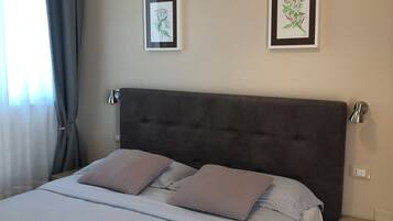 Una cama bien hecha con cabecera gris, dos mesitas de noche con lámparas y dos impresiones botánicas enmarcadas en la pared.