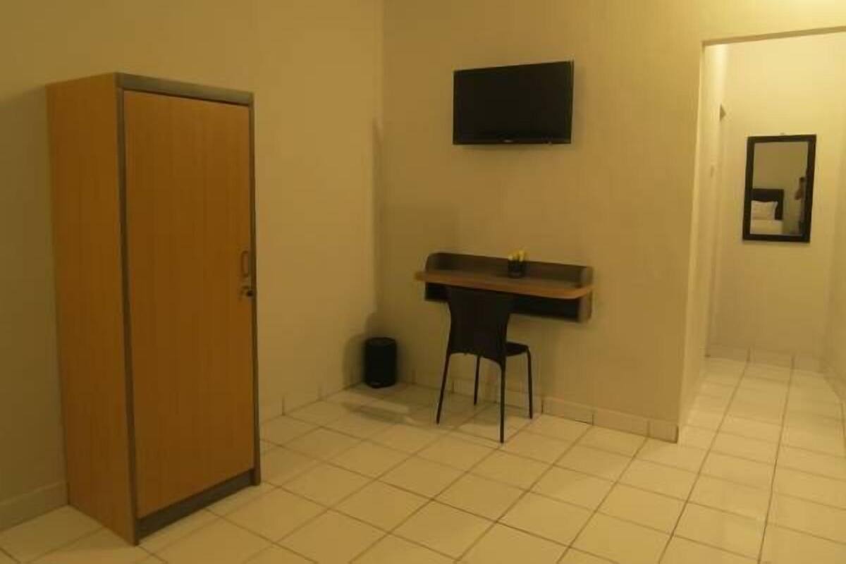 A small room with a wooden wardrobe, a small desk with a chair, a television mounted on the wall, and a mirror on the wall.