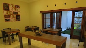 Un área de comedor con varias mesas y sillas, una mesa central decorada con un arreglo floral, y un ventanal con vistas a la vegetación exterior.