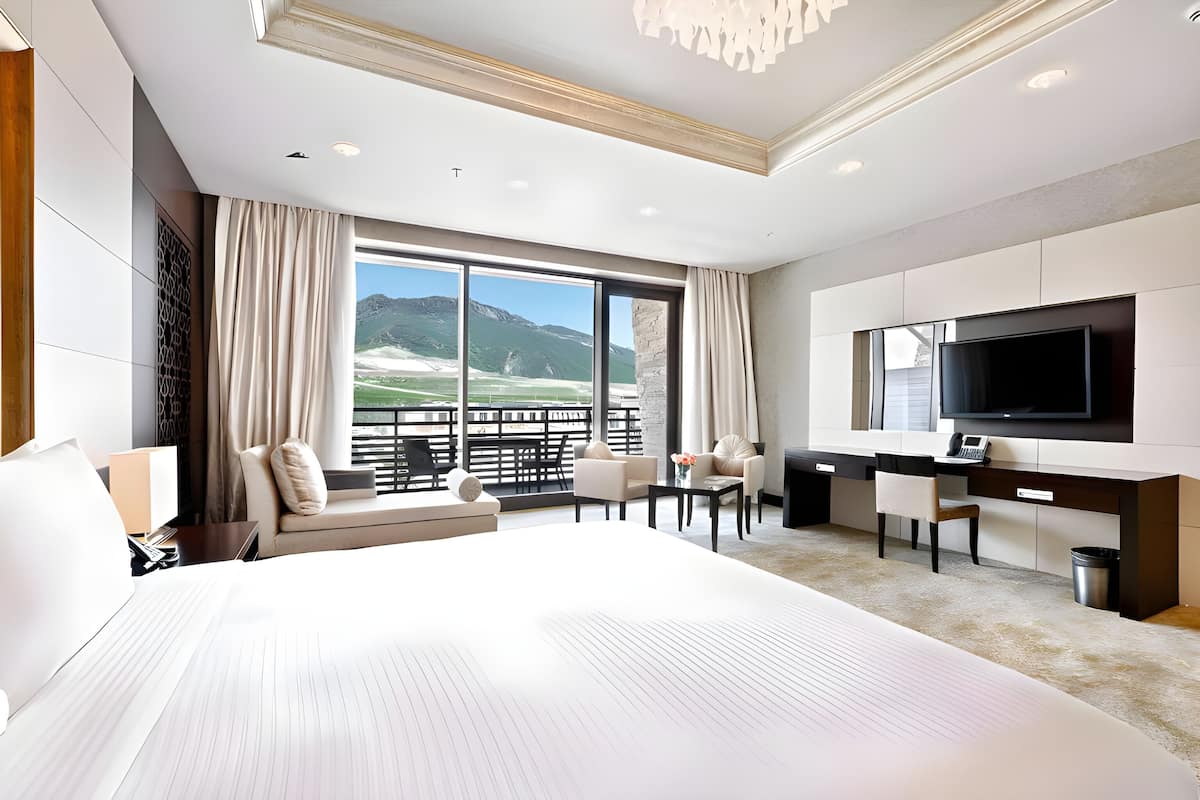A modern hotel room with a large bed, a desk, a TV, and a view of mountains.