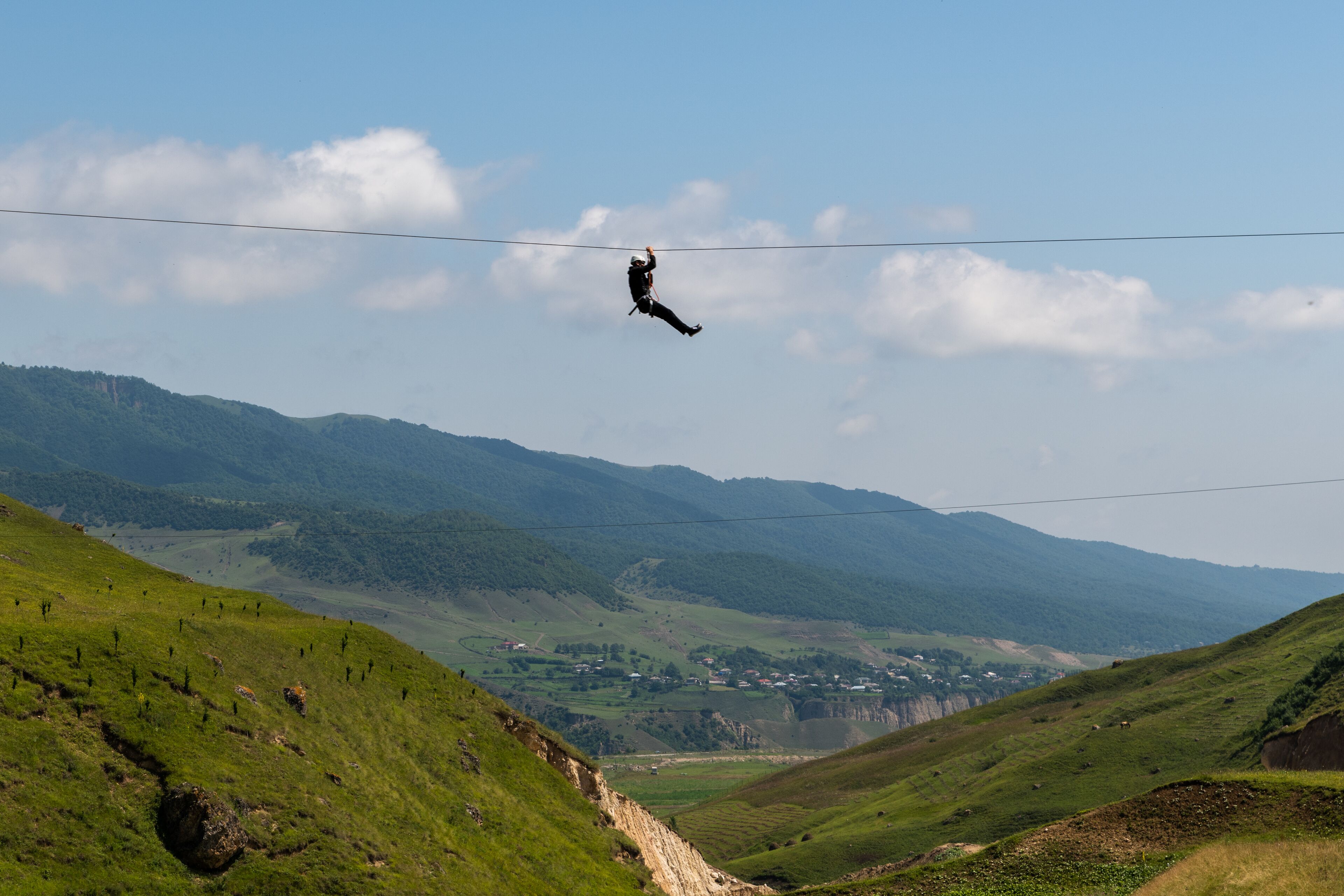 Eine Person springt mit einem Bungee-Seil von einem hohen Drahtseil über ein Tal mit grünen Hügeln und einer in der Ferne liegenden Stadt.