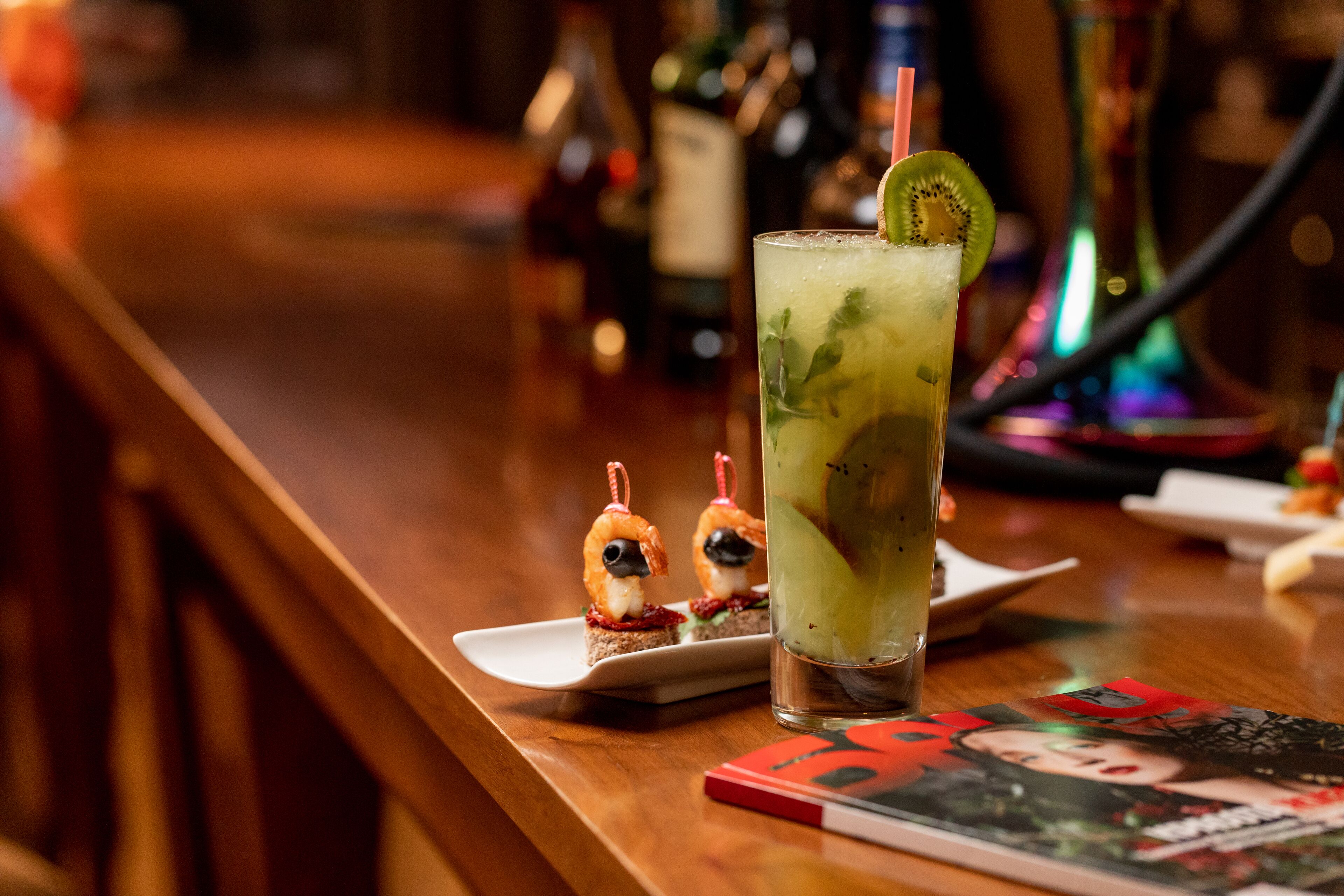 Un bar, avec un verre d’un cocktail vert décoré d’une tranche de kiwi, une assiette contenant deux brochettes de crevettes et un magazine posé sur la table.