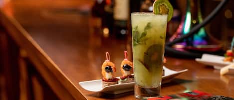 Un bar, avec un verre d’un cocktail vert décoré d’une tranche de kiwi, une assiette contenant deux brochettes de crevettes et un magazine posé sur la table.