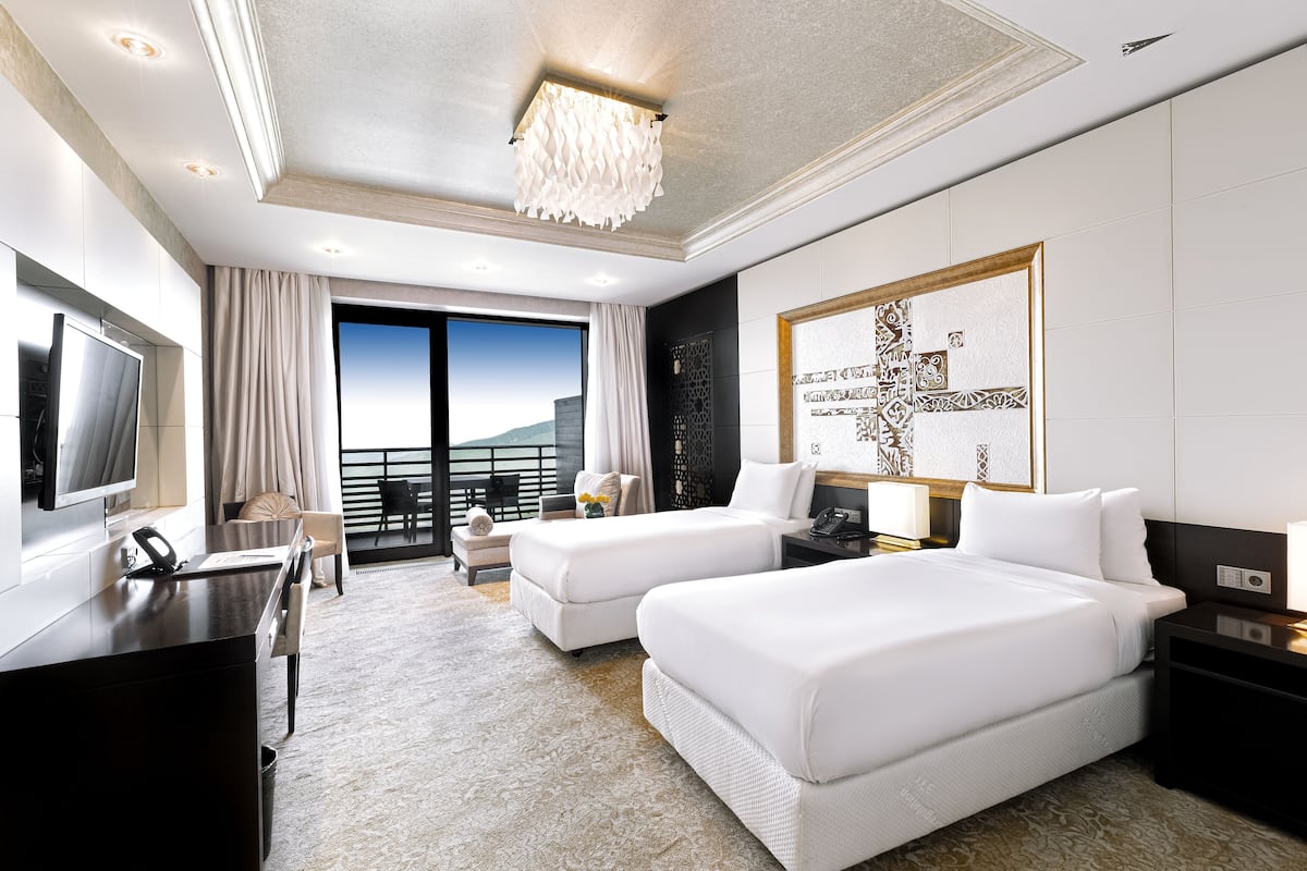 A modern hotel room with two beds, a desk, a TV, and a balcony with a view.