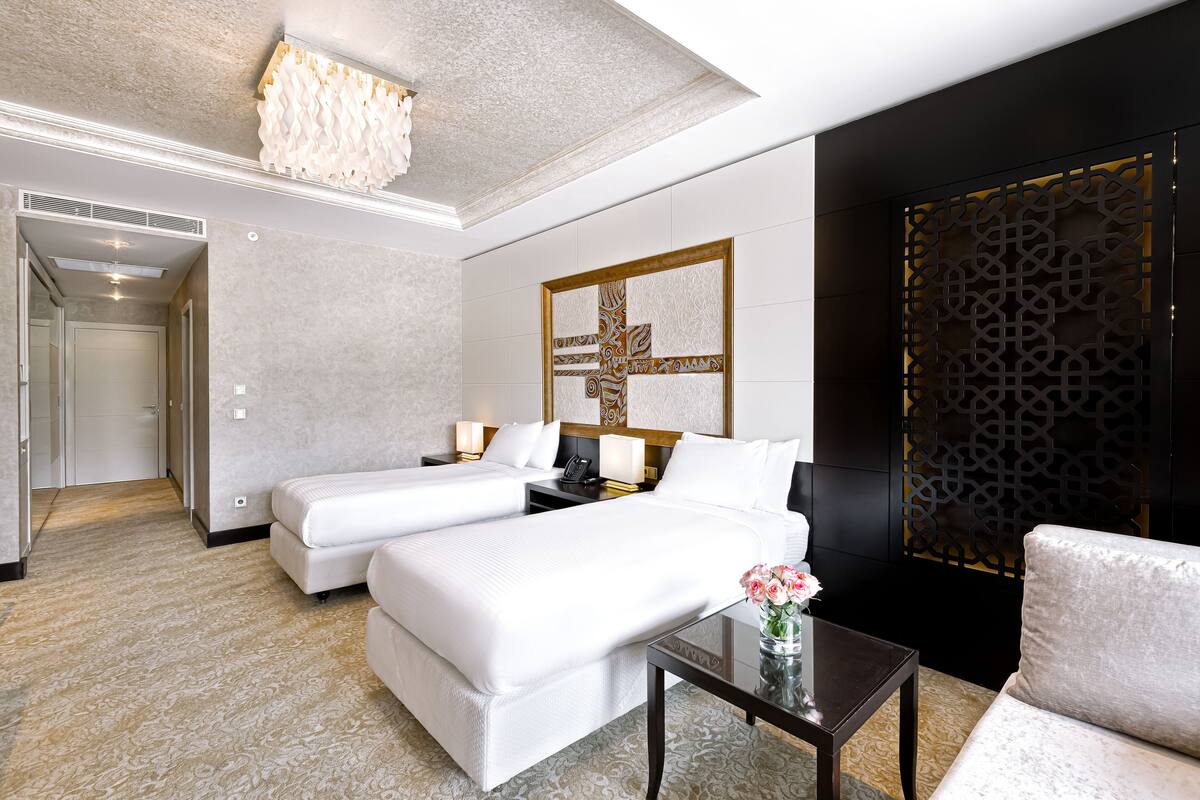 A hotel room with two beds, a sitting area with a table and chair, a decorative wall piece, and a patterned carpet.