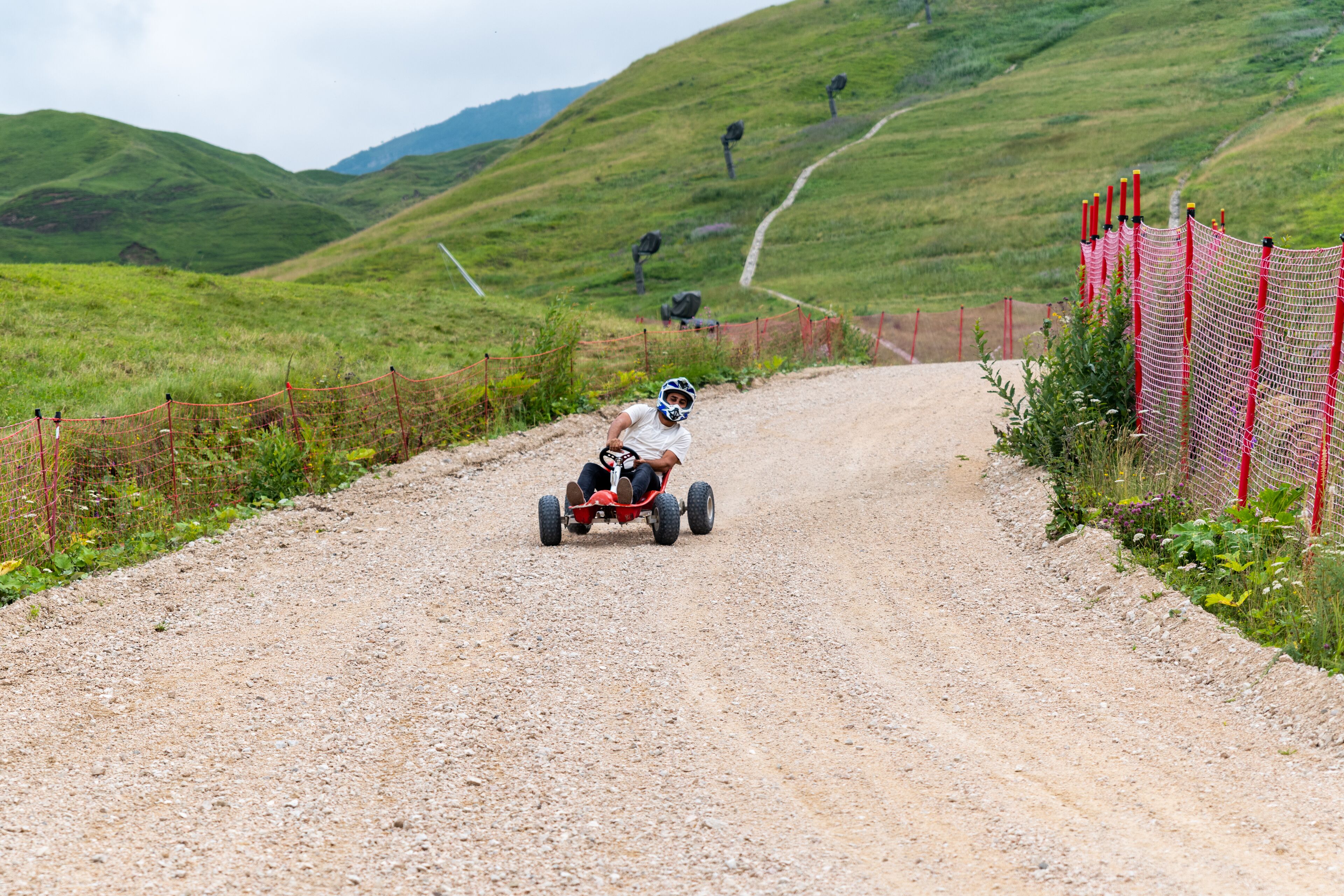 Iemand rijdt op een quad over een grindweg, met een rode afrastering aan de rechterkant en groene heuvels op de achtergrond.