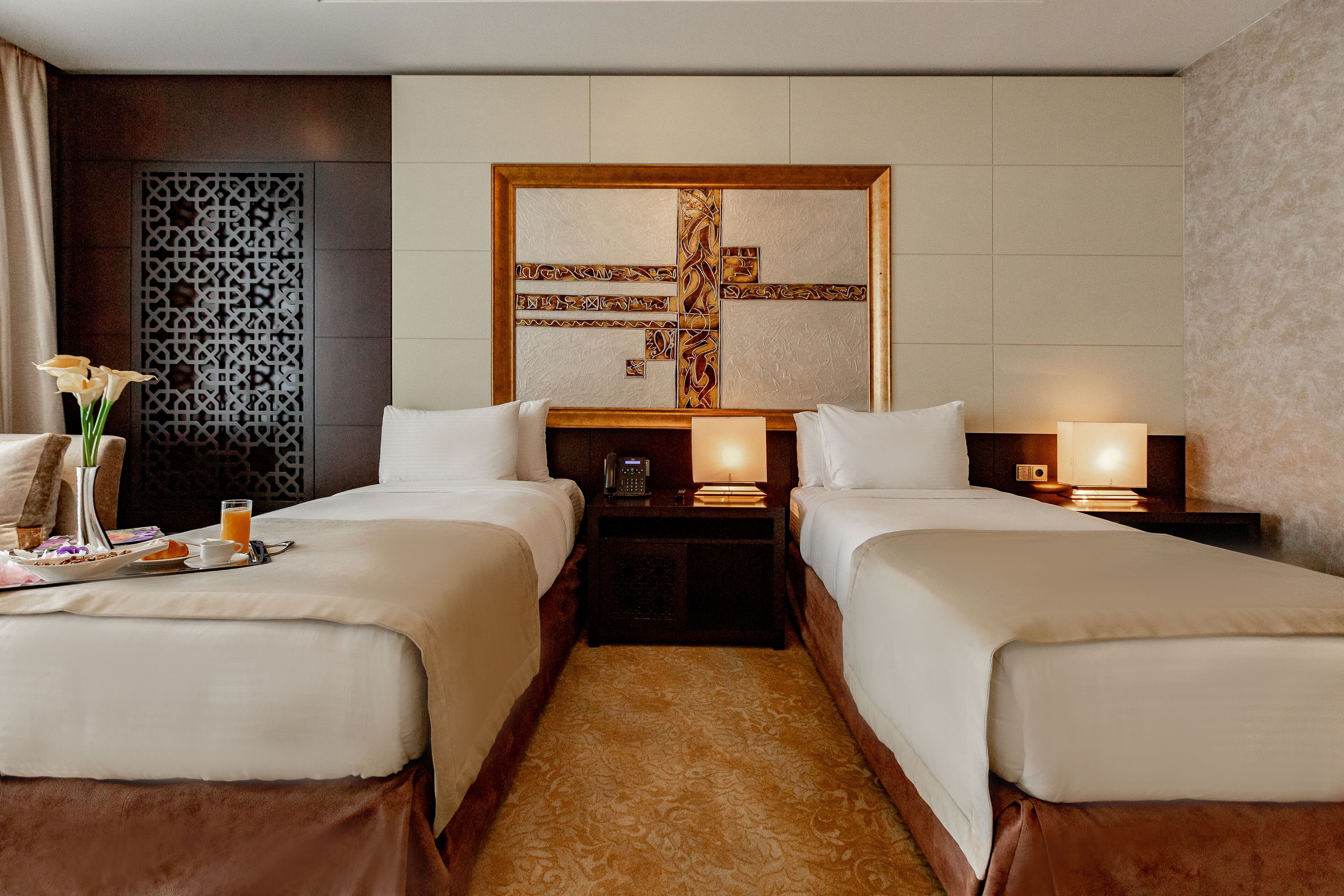 A hotel room with two beds, a decorative wall panel, a bedside table with a lamp, and a tray with breakfast items.